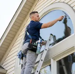 Professional window cleaner on a ladder using a squeegee to clean an exterior arched window, Bakersfield, CA.
